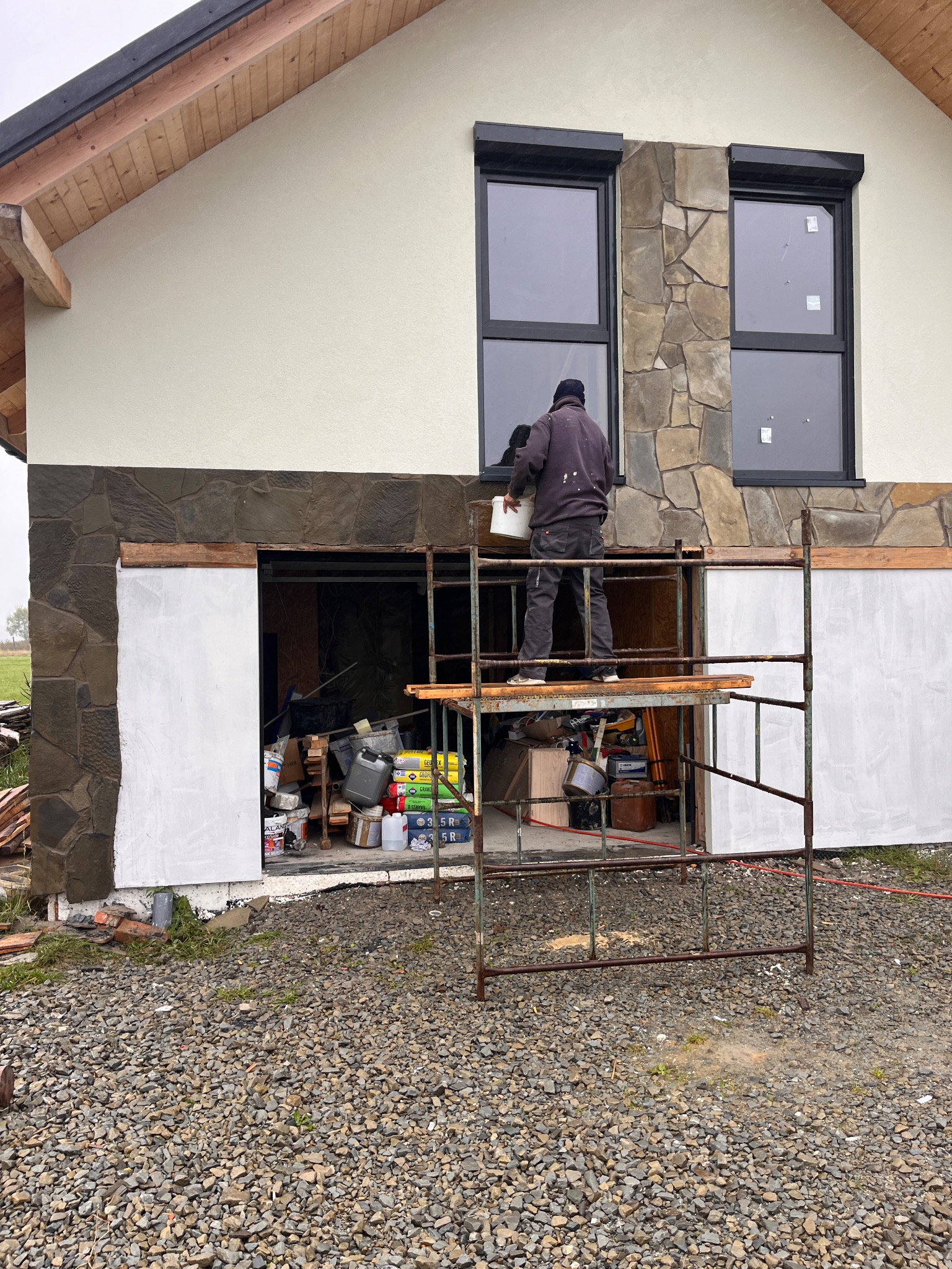 Pracownik na rusztowaniu maluje elewację domu z kamiennymi elementami. Widoczne okna z roletami zewnętrznymi i materiały budowlane w otwartym garażu.