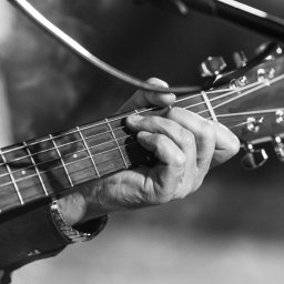 Laboratorium i Studio Fotograficzne RAW S.C. - Czarno-białe zbliżenie dłoni grającej akord na gitarze akustycznej, widoczny gryf, struny i główka gitary, oświetlone światłem reflektorów, z mikrofonem w tle.