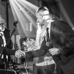 Laboratorium i Studio Fotograficzne RAW S.C. - Czarno-białe zdjęcie uśmiechniętych mężczyzn w garniturach, ściskających dłonie na tle kobiety z bukietem i kościelnych dekoracji, oświetlonych promieniami światła.