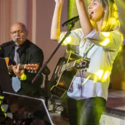 Wokalistka z gitarą śpiewa na scenie, oświetlona żółtym światłem. W tle muzyk gra na gitarze. Widoczny statyw z nutami. Ujęcie z dołu, podkreślające energię występu.