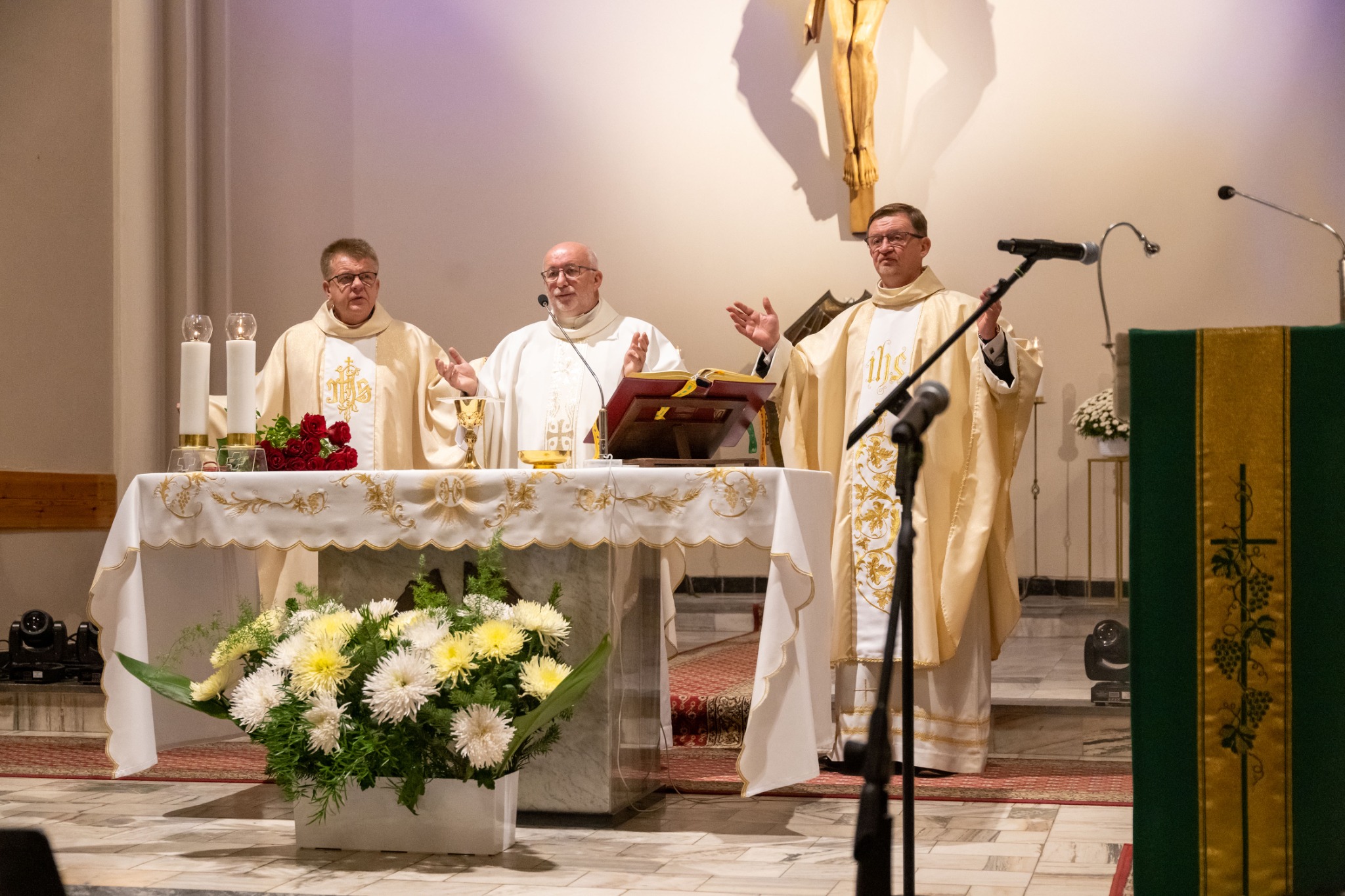 Trzech księży w szatach liturgicznych celebruje mszę przy ołtarzu udekorowanym kwiatami i świecami, pod krzyżem w kościele.