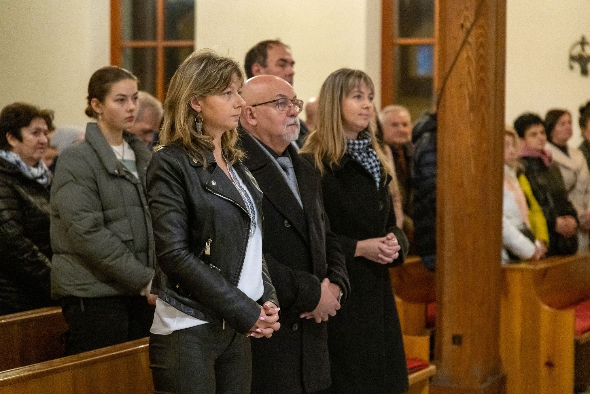 Uczestnicy uroczystości religijnej w kościele w Pniewach. Grupa osób stoi w ławkach, widoczne drewniane elementy wystroju wnętrza i okna.