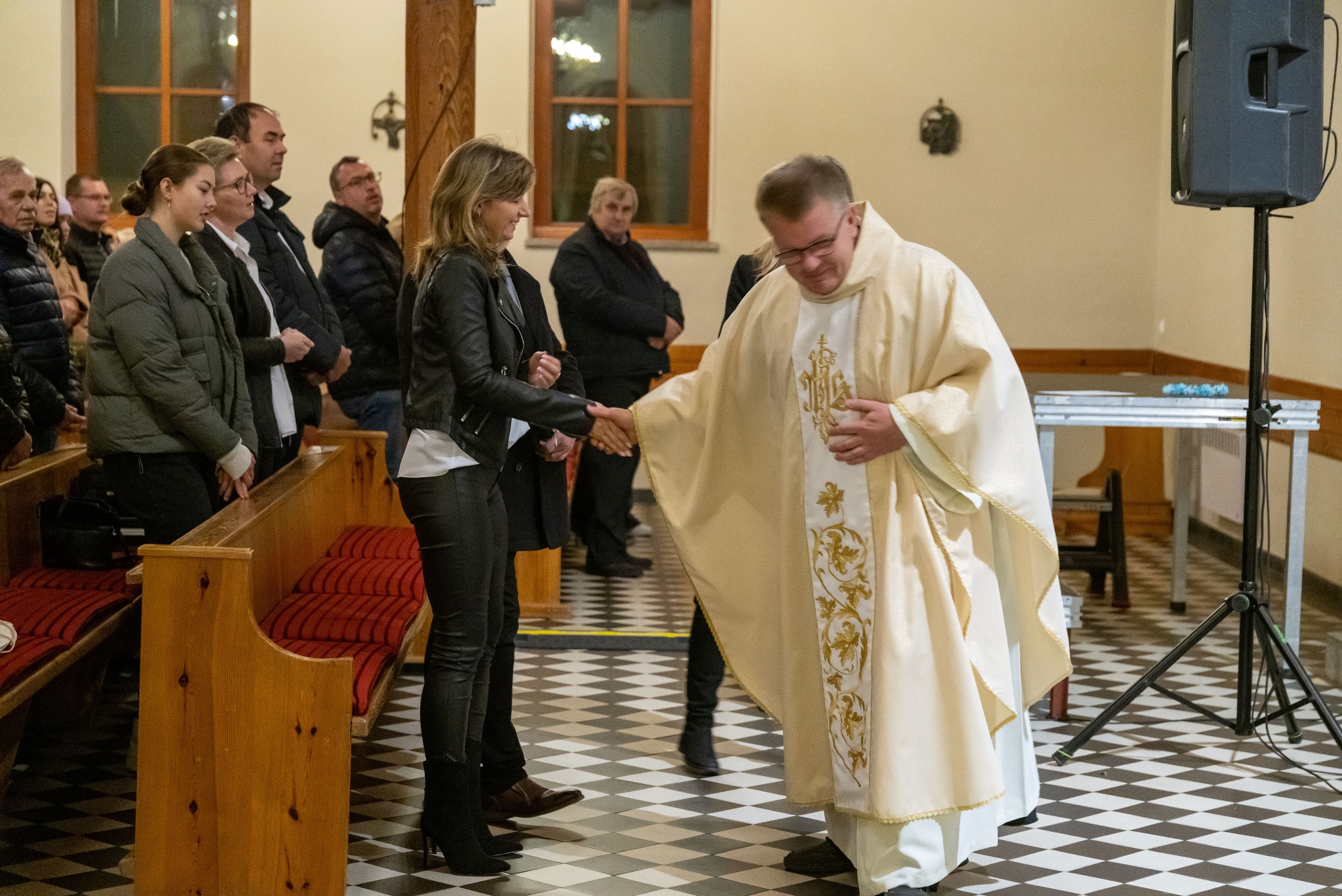 Ksiądz w szatach liturgicznych w kościele wita się z kobietą na tle wiernych i wzorzystej posadzki. Ujęcie z boku, z widocznymi elementami wyposażenia kościoła.