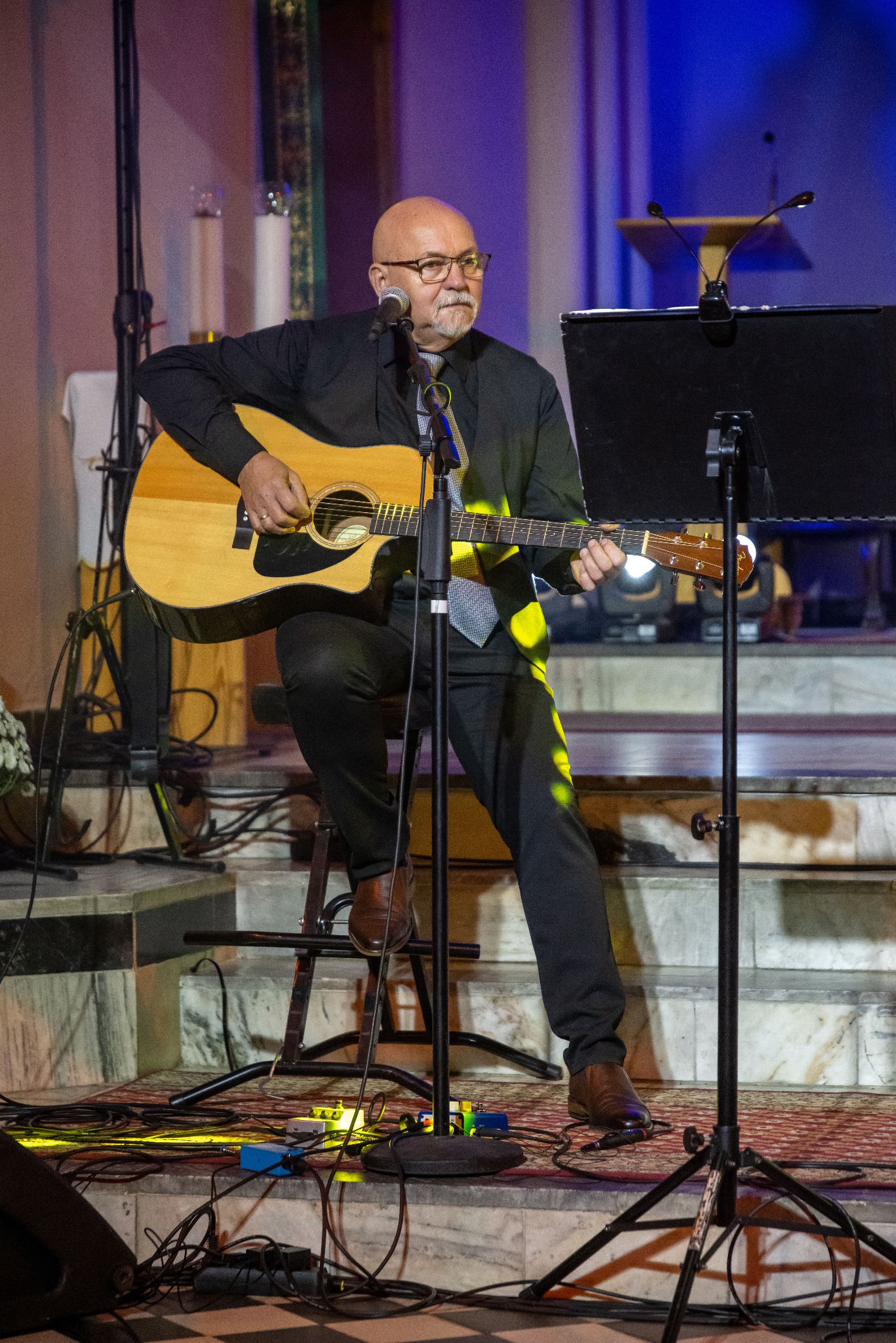 Mężczyzna w okularach gra na gitarze akustycznej na scenie, siedząc na stołku, z mikrofonem i statywem na nuty. W tle widać elementy wystroju kościelnego.