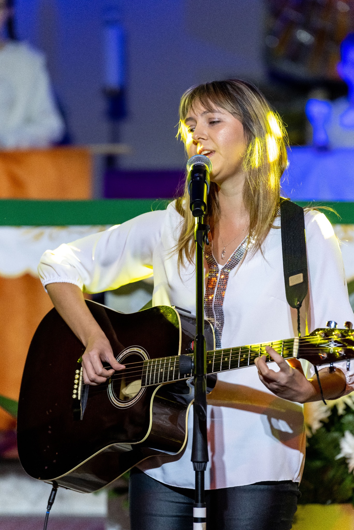 Kobieta śpiewa do mikrofonu, grając na gitarze akustycznej. Scena oświetlona ciepłym światłem, w tle dekoracje. Ubrana w białą bluzkę z ozdobnymi cekinami.