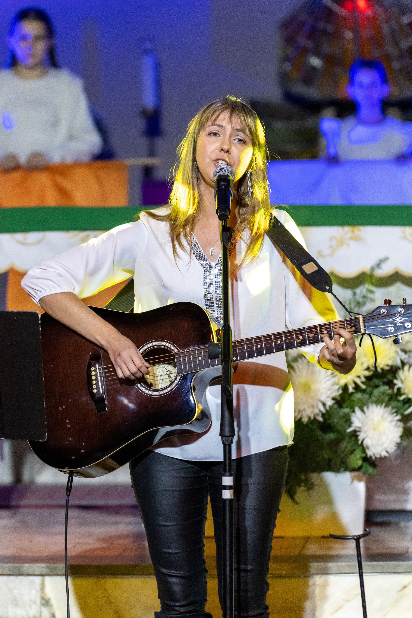 Kobieta z gitarą akustyczną śpiewa do mikrofonu na scenie, w tle dekoracje i osoby. Scena oświetlona światłem reflektorów, widoczne kwiaty i elementy dekoracyjne.