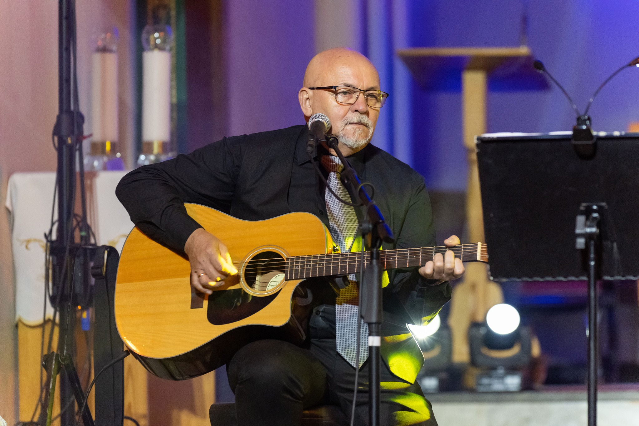 Mężczyzna w okularach i koszuli gra na gitarze akustycznej na scenie z mikrofonami i oświetleniem. W tle widoczne elementy dekoracyjne.
