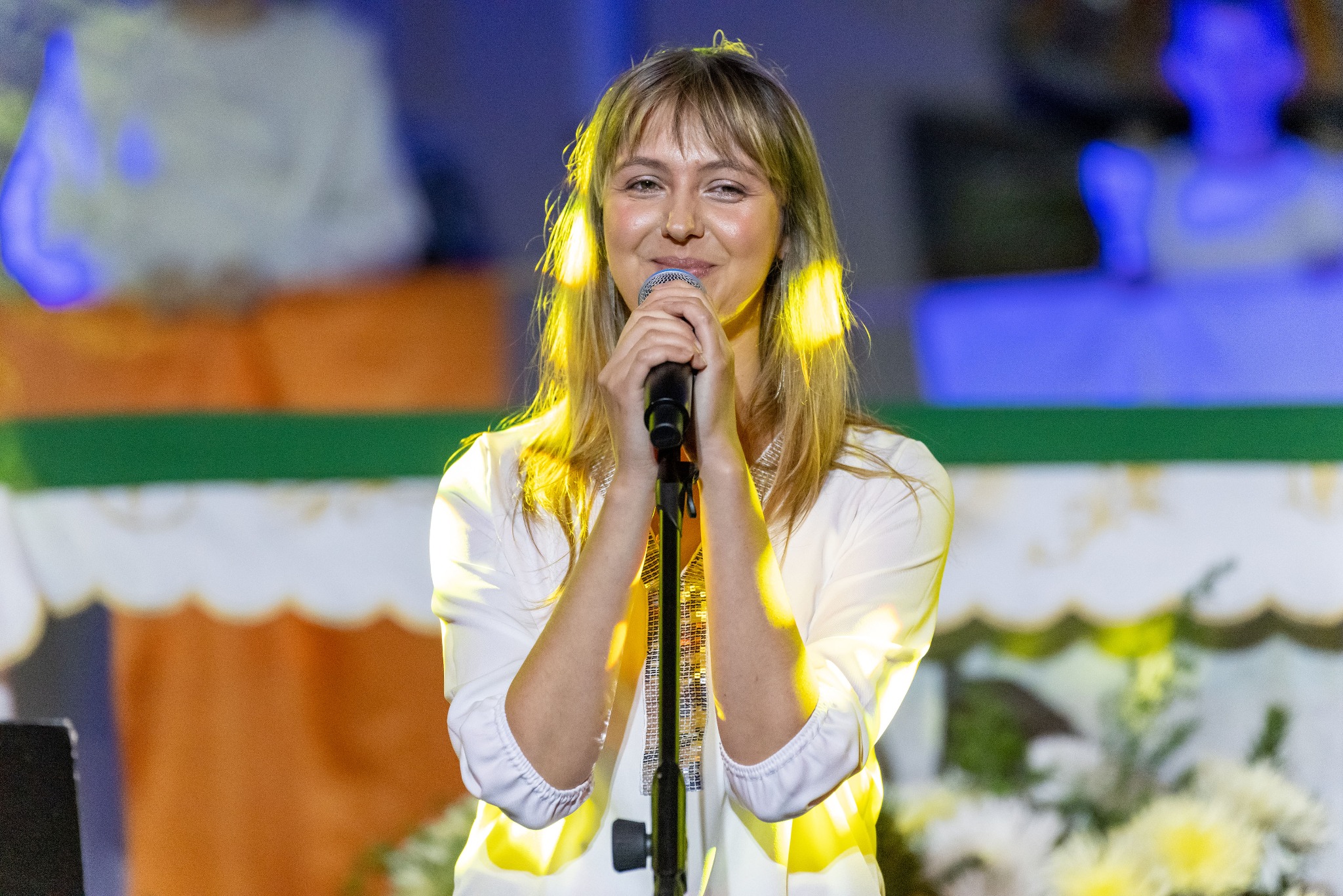 Uśmiechnięta blondynka w białej bluzce śpiewa do mikrofonu na scenie, w tle dekoracje i publiczność. Scena oświetlona reflektorami.