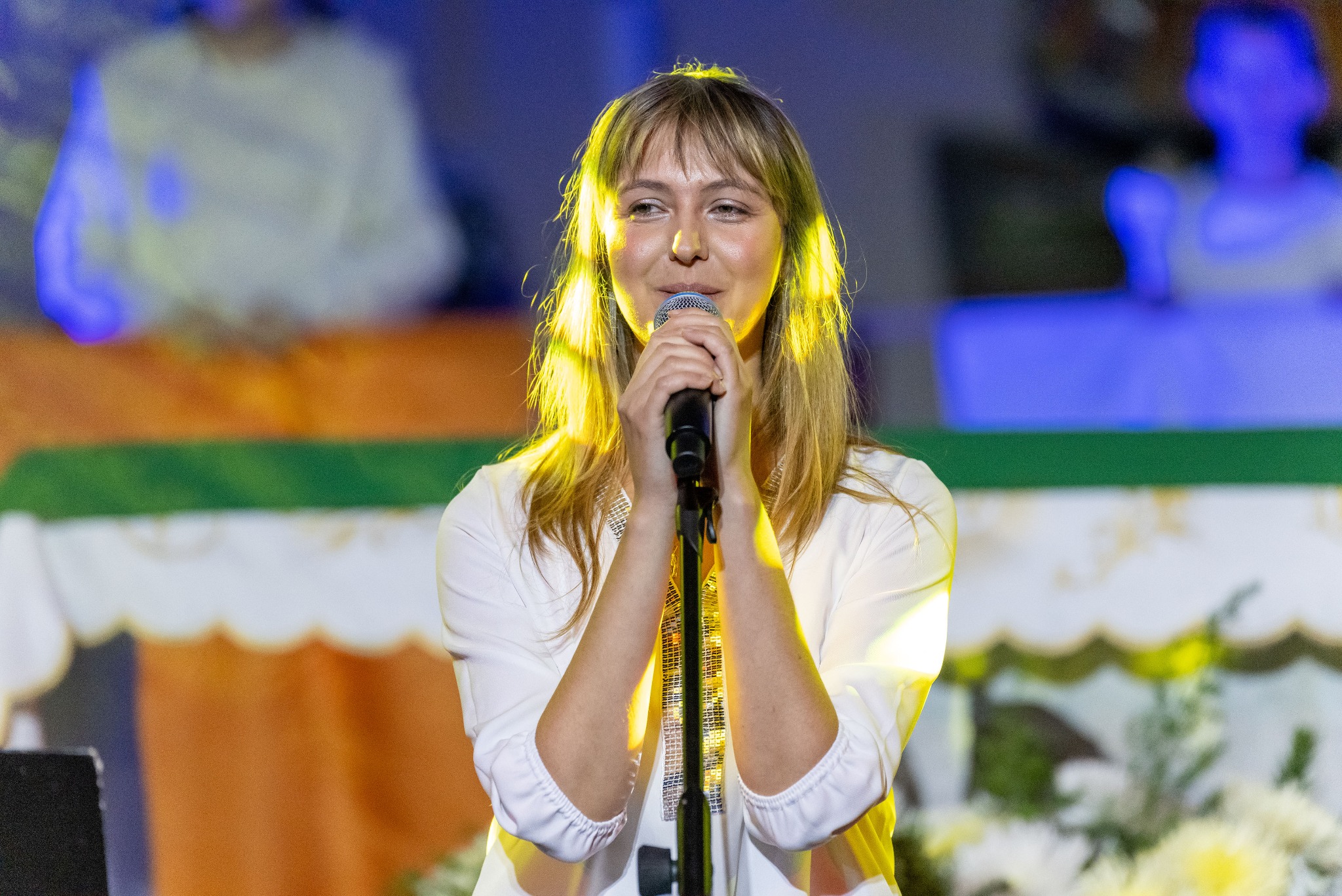 Kobieta z mikrofonem, na scenie w białej bluzce z ozdobami, oświetlona ciepłym światłem. W tle dekoracje i publiczność.