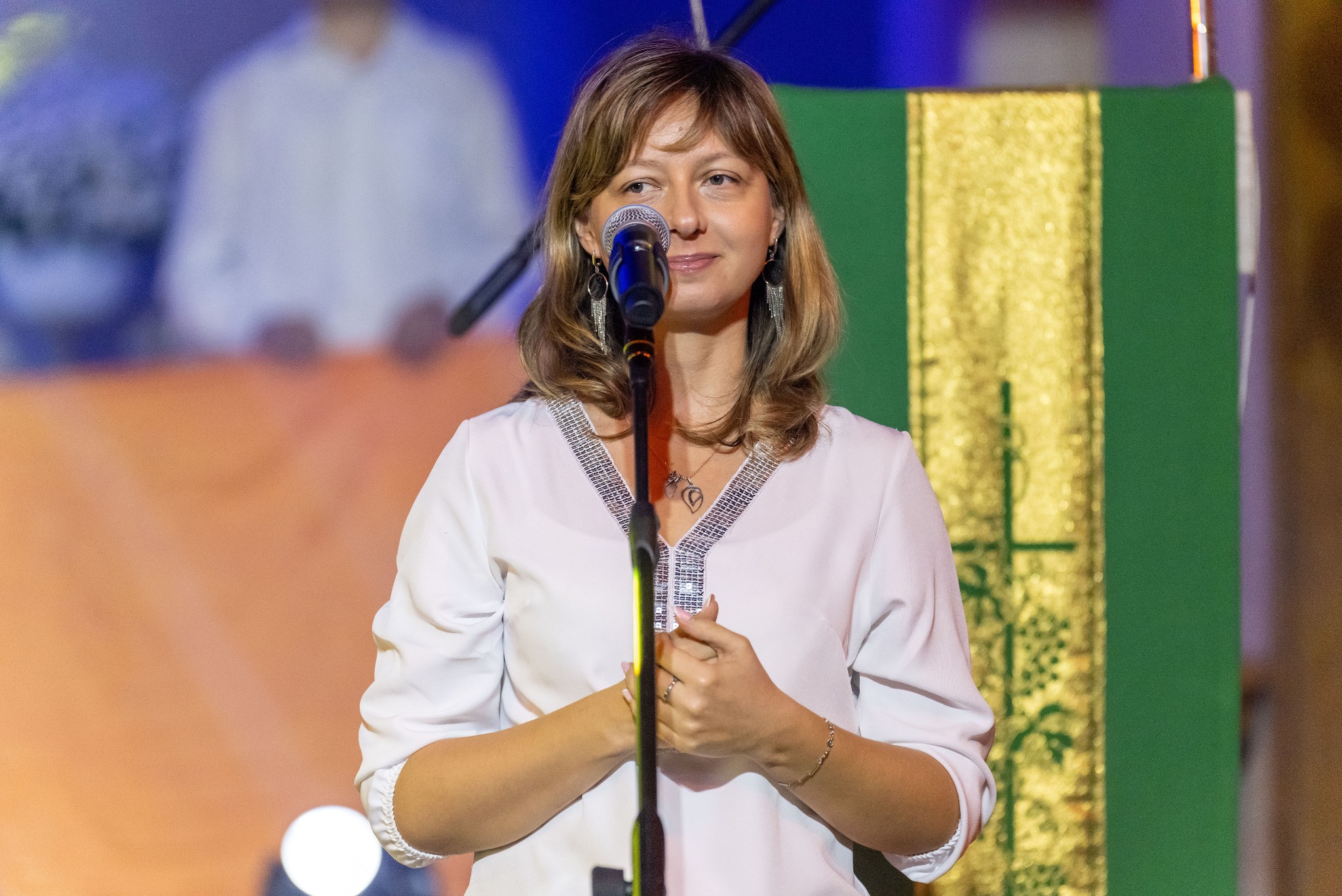 Kobieta z mikrofonem na scenie, w białej bluzce z ozdobnym dekoltem, z delikatnym uśmiechem. W tle dekoracja w odcieniach złota i zieleni, oświetlenie sceniczne.