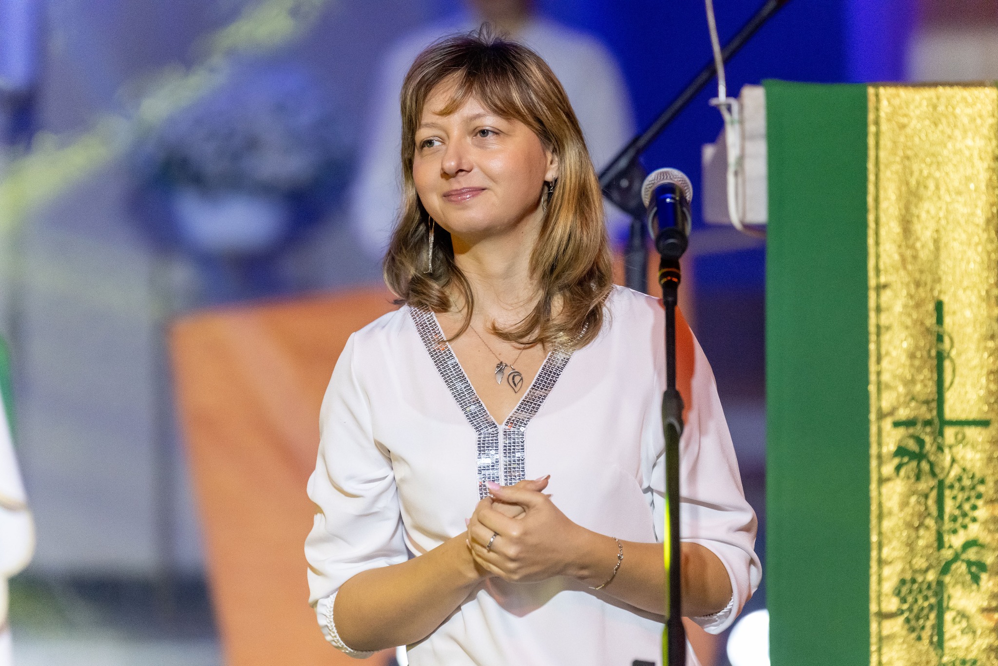 Portret kobiety w białej bluzce z błyszczącymi aplikacjami, stojącej na scenie obok mikrofonu i dekoracji. Uśmiech, subtelny makijaż i biżuteria.