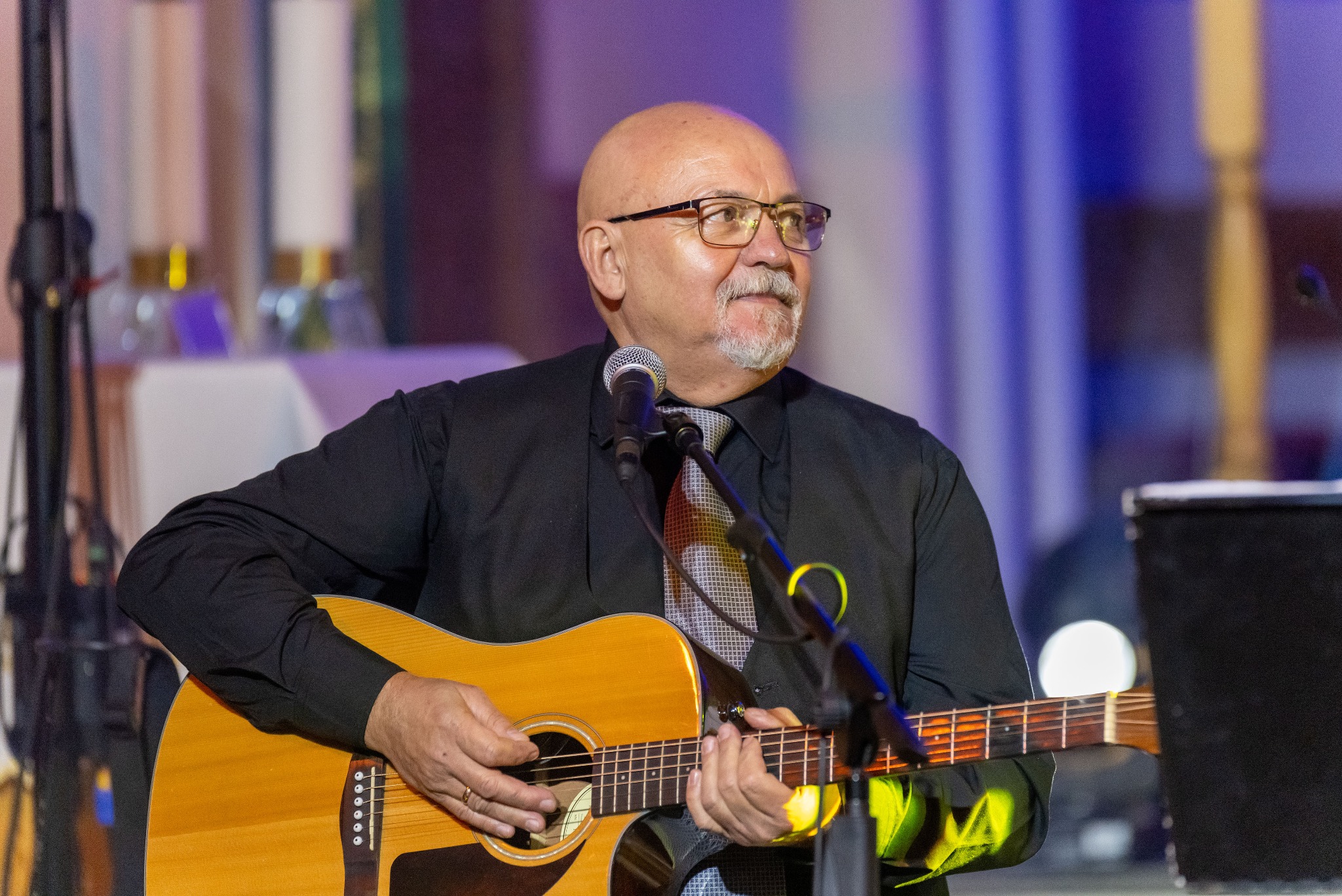 Mężczyzna w okularach gra na gitarze akustycznej na scenie, ubrany w czarną koszulę i krawat. Widoczny mikrofon i oświetlenie sceniczne w tle.