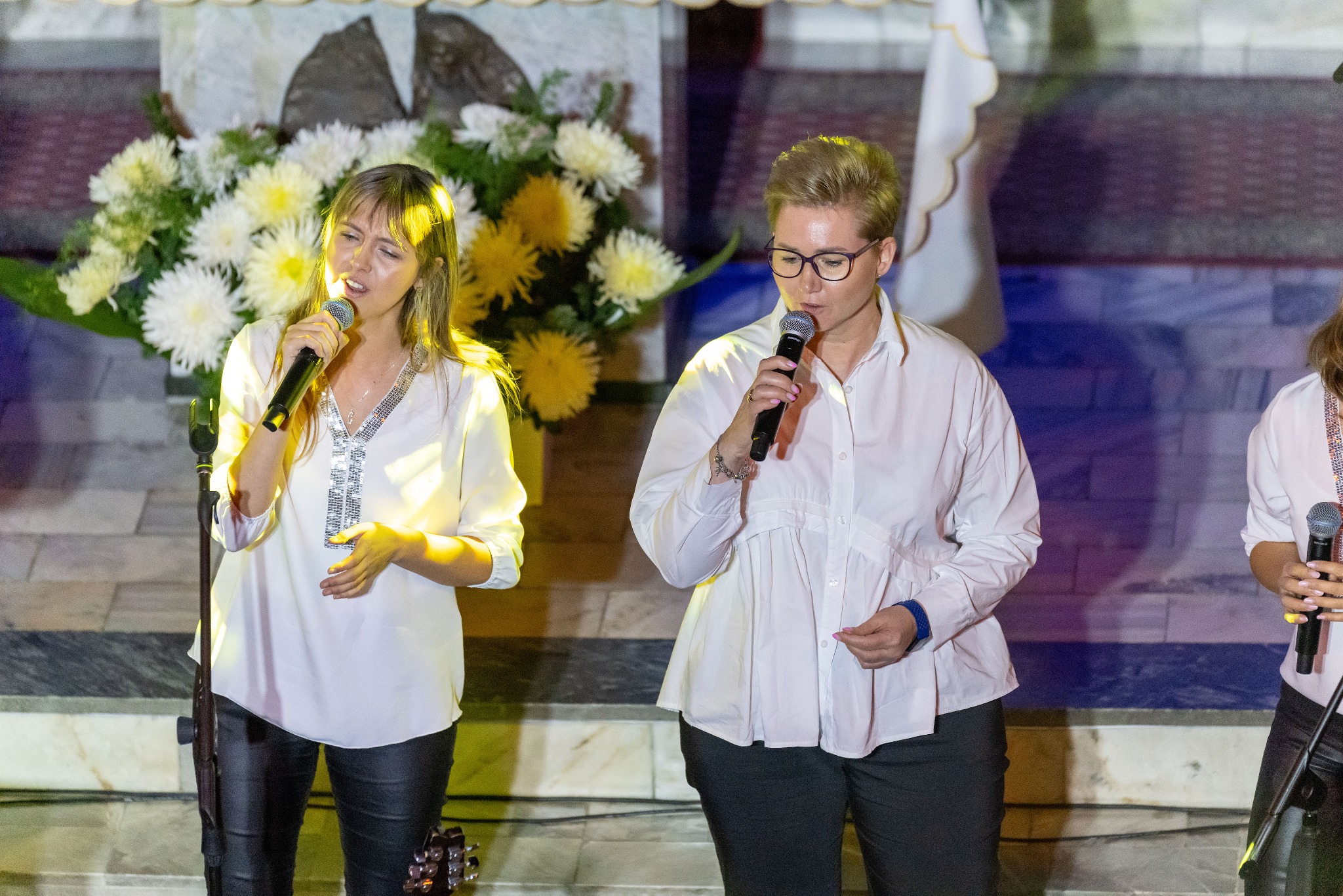 Dwie kobiety śpiewają do mikrofonów na scenie z kwiatami w tle. Jedna w okularach, obie w białych bluzkach. Widoczny fragment gitary. Scena oświetlona.