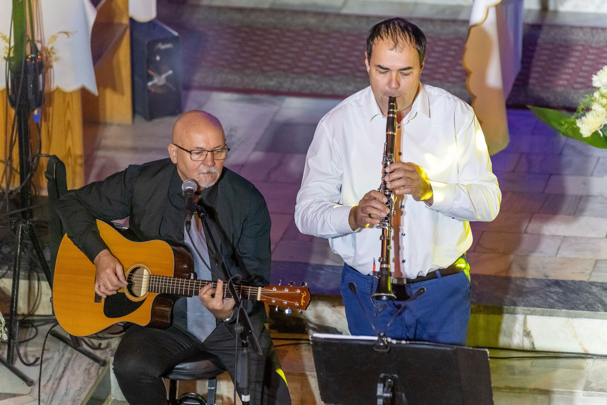 Duet muzyczny: gitarzysta w czarnej marynarce i klarnecista w białej koszuli występują na tle kościoła.