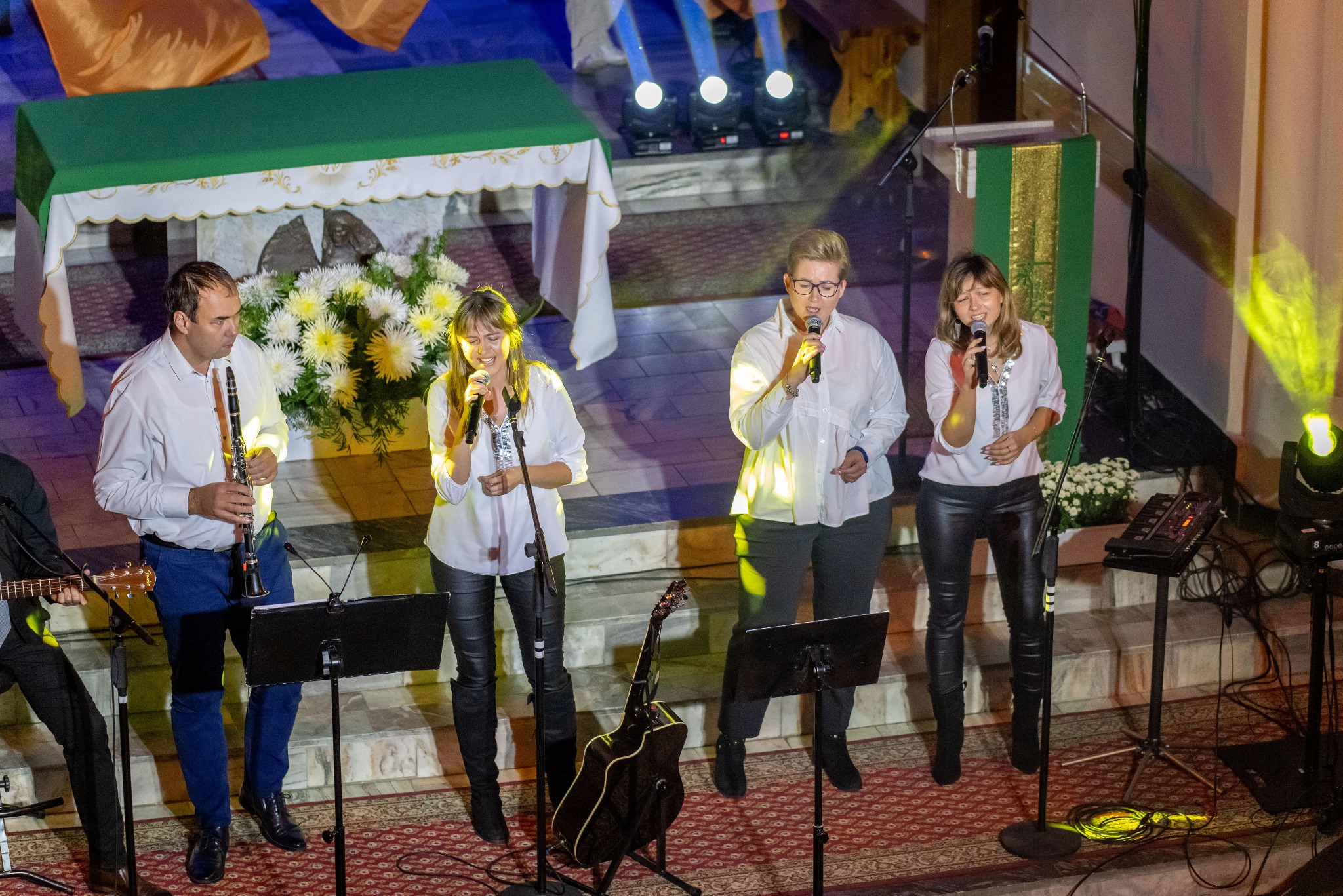 Zespół muzyczny z wokalistkami, klarnecistą i gitarzystą występuje na scenie w kościele, oświetlony reflektorami, z kwiatami w tle.