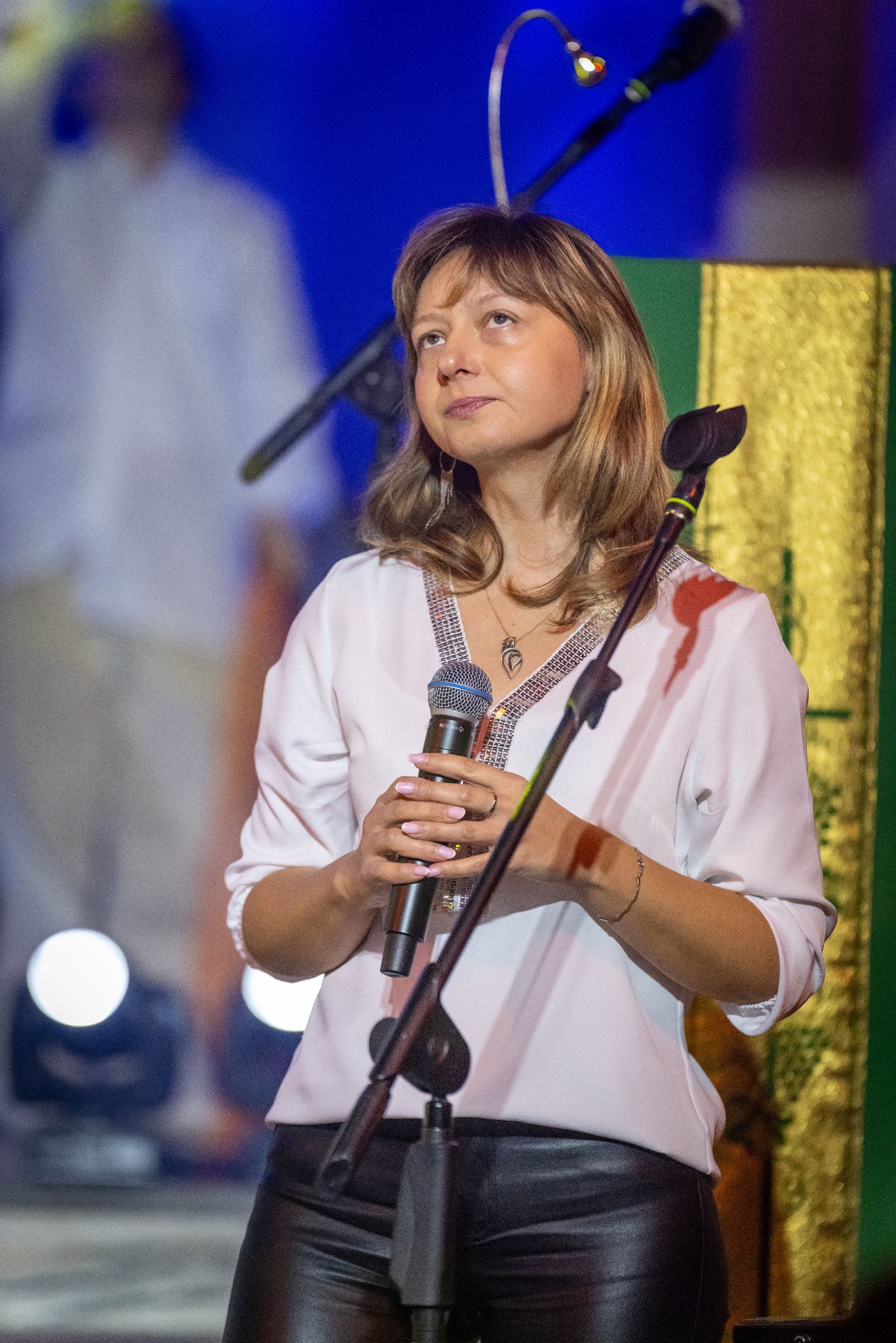 Kobieta z mikrofonem Beta 58A na scenie, w białej bluzce z cekinami i czarnych spodniach. Reflektory tworzą jasne punkty w tle.