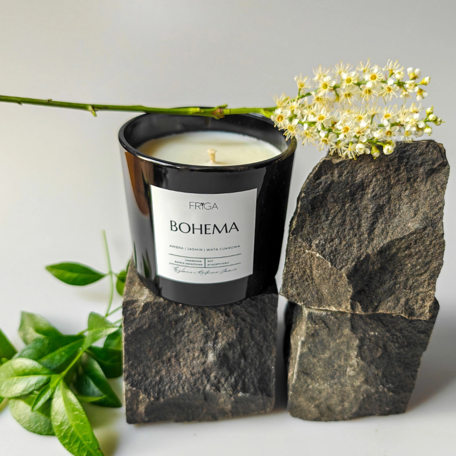 Czarna świeca zapachowa Friga Bohema z nutami ambra, jaśminu i waty cukrowej, umieszczona na kamiennych blokach, ozdobiona kwiatami i zielonymi liśćmi.