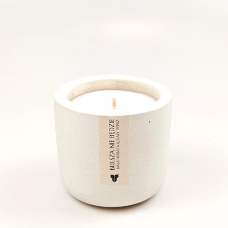 Minimalistyczna, biała świeca w betonowym pojemniku z etykietą 'Bielsza nie będzie' o zapachu białej herbaty i białego pieprzu, na jasnym tle.