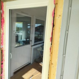 Fast Bygg - Montaż okna balkonowego: widoczne ocieplenie pianką montażową i wełną mineralną. W tle drewniany taras i wnętrze domu. Jasne światło dzienne.