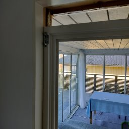 Fast Bygg - Wnętrze w trakcie wykończenia: widok przez białe futryny na przeszkloną werandę z drewnianą podłogą i stołem przykrytym błękitnym obrusem. Częściowo odsłonięta konstrukcja sufitu.