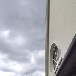 Fast Bygg - Fragment elewacji budynku z okrągłym oknem i widocznym niebem. Ujęcie z dołu podkreśla architekturę i specyficzny kształt okna. Minimalistyczny design budynku.