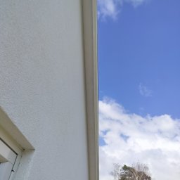 Fast Bygg - Biała elewacja domu z widocznym oknem i fragmentem dachu na tle błękitnego nieba z chmurami i drzewami, ujęcie z dołu.