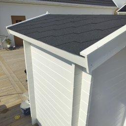 Fast Bygg - Biała, drewniana altana z czarnym, bitumicznym dachem na drewnianym tarasie. Widoczne ślady farby i wiadro z farbą, sugerujące świeże malowanie.