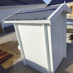 Fast Bygg - Biała, drewniana altanka z szarym dachem na drewnianym tarasie, widziana z góry. W tle domy z czarną dachówką i oknami dachowymi.