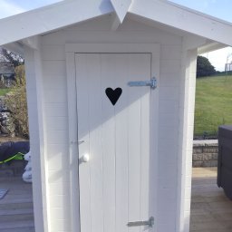 Fast Bygg - Biała, drewniana altanka z sercem wyciętym w drzwiach, widoczna na tle zielonego trawnika i błękitnego nieba. Detale: metalowe zawiasy, drewniana podłoga.