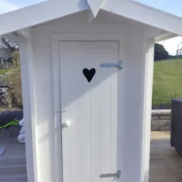 Biała, drewniana altanka z sercem wyciętym w drzwiach, widoczna na tle zielonego trawnika i błękitnego nieba. Detale: metalowe zawiasy, drewniana podłoga.