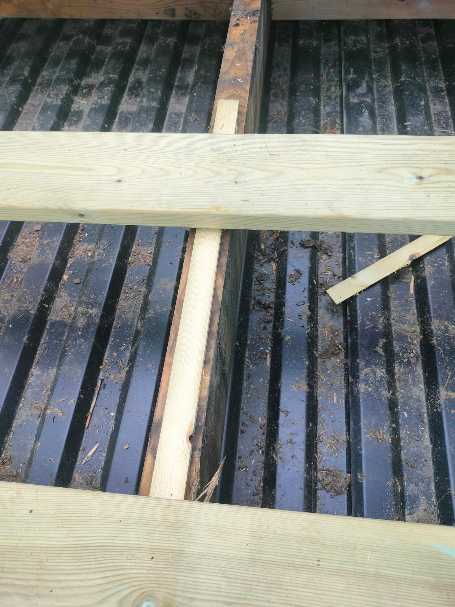 Konstrukcja drewniana na czarnej, ryflowanej powierzchni. Widoczne elementy montażowe i fragmenty drewna. Budowa w toku, z bliska, z góry.