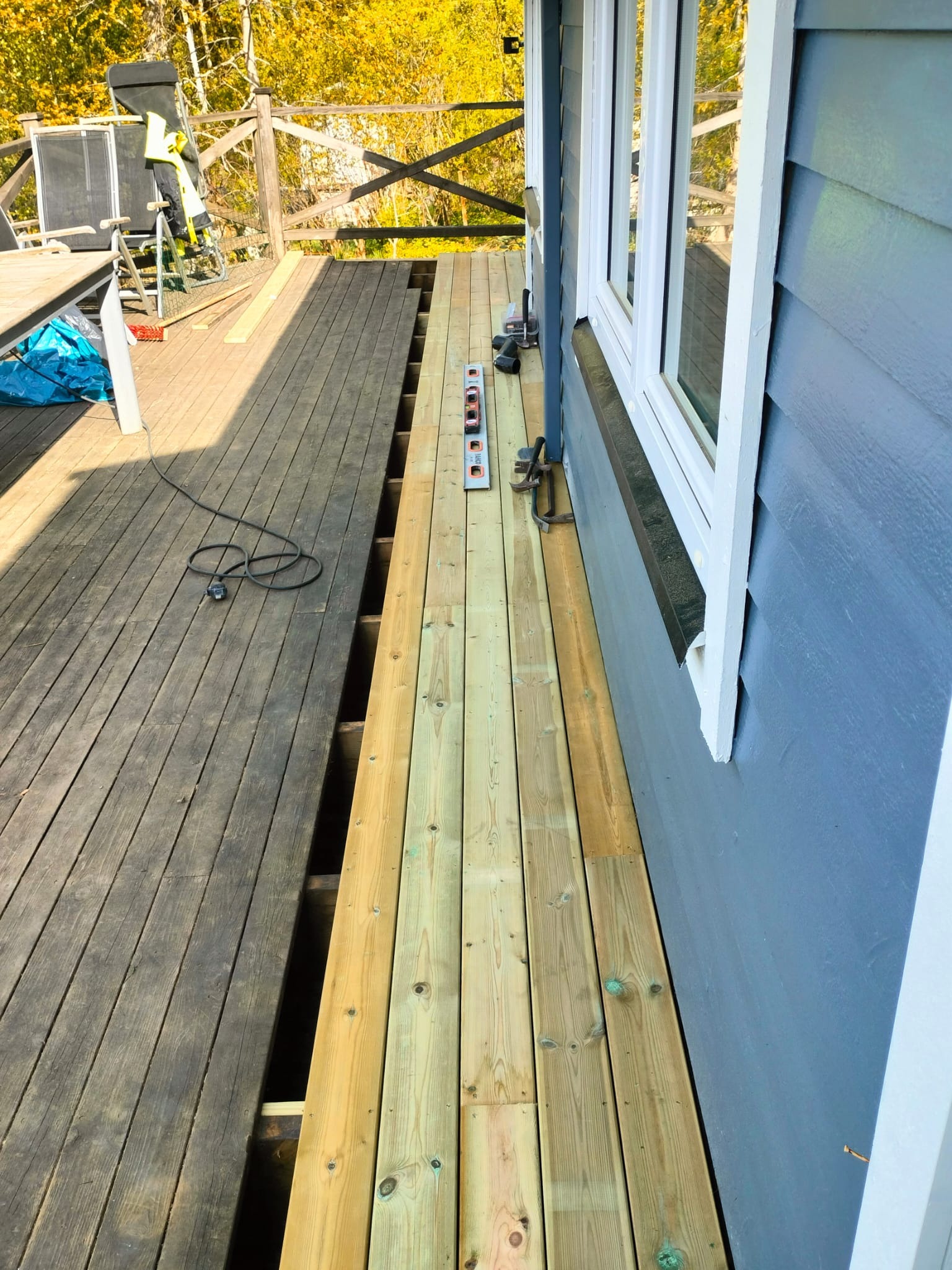 Montaż desek tarasowych: część starego, ciemnego tarasu obok nowej, jasnej części. Widoczna poziomica i narzędzia, ściana domu z oknem w tle.