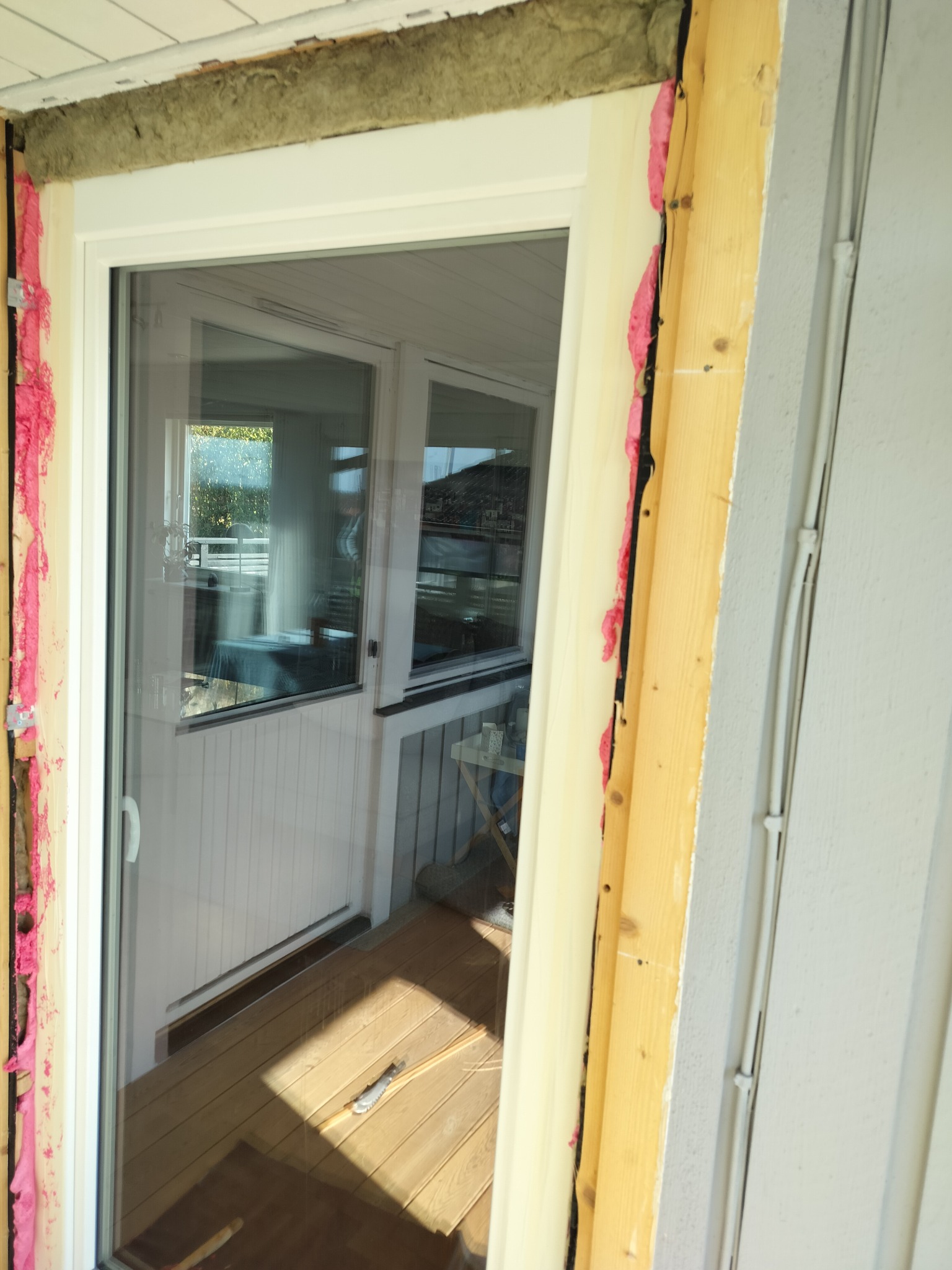 Montaż okna balkonowego: widoczne ocieplenie pianką montażową i wełną mineralną. W tle drewniany taras i wnętrze domu. Jasne światło dzienne.