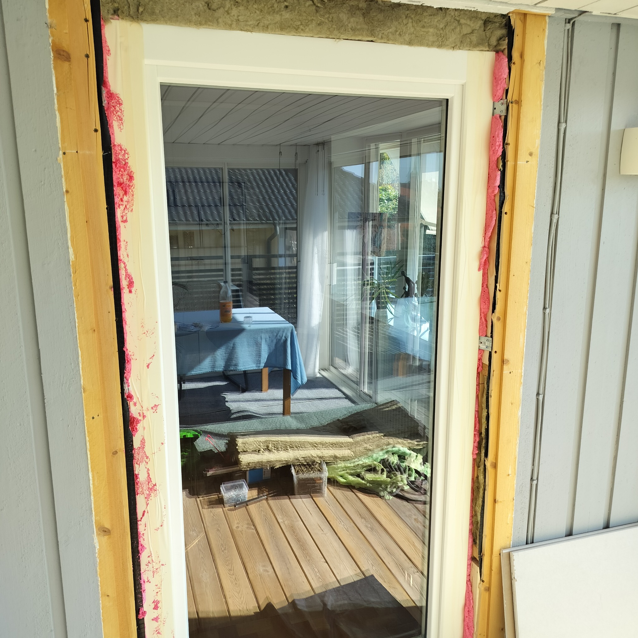 Montaż okna: widoczne ocieplenie, pianka montażowa i drewniane elementy ramy. W tle taras z deskami i widok na wnętrze z niebieskim obrusem.