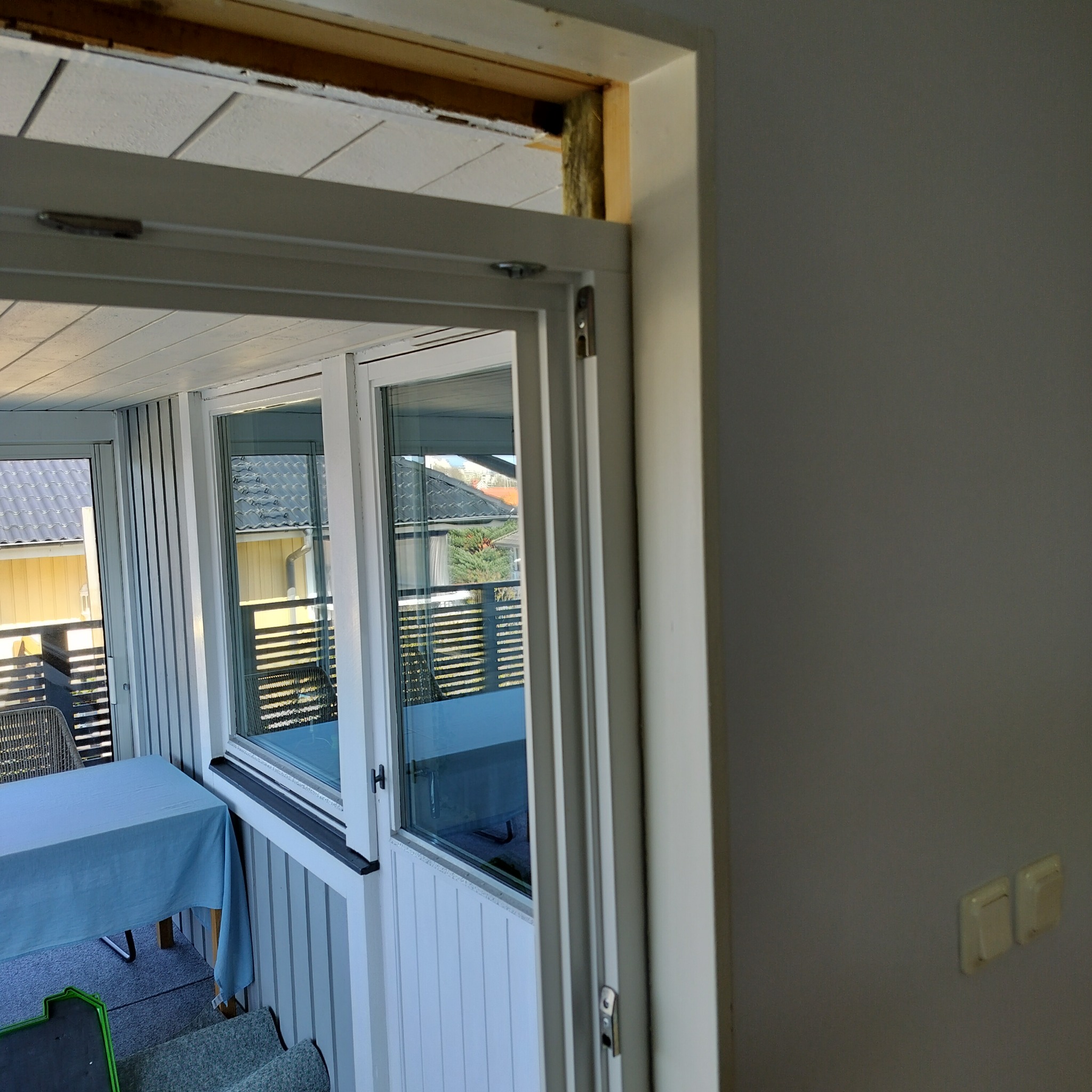 Otwór drzwiowy w trakcie prac wykończeniowych, widoczna izolacja w nadprożu, fragment balkonu z oknami i stolikiem w tle. Detal wnętrza w trakcie remontu.