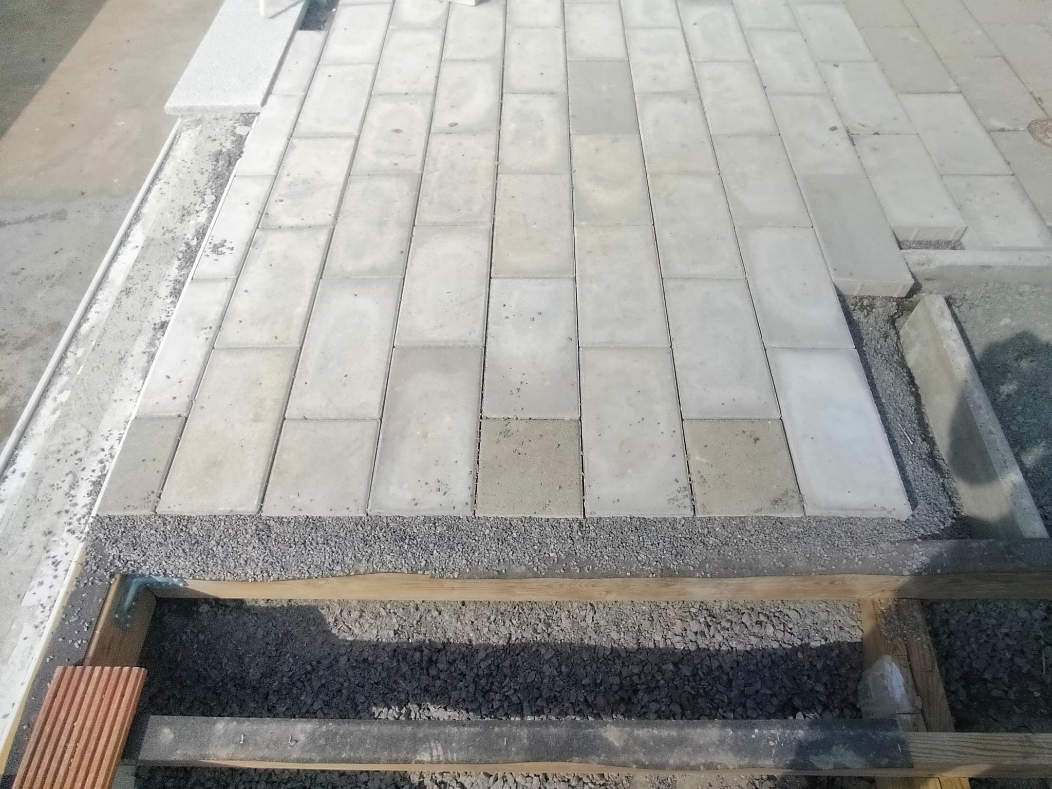 Budowa tarasu: ułożona kostka brukowa na podsypce z kruszywa, widoczne drewniane elementy konstrukcyjne. Etap prac w słoneczny dzień.