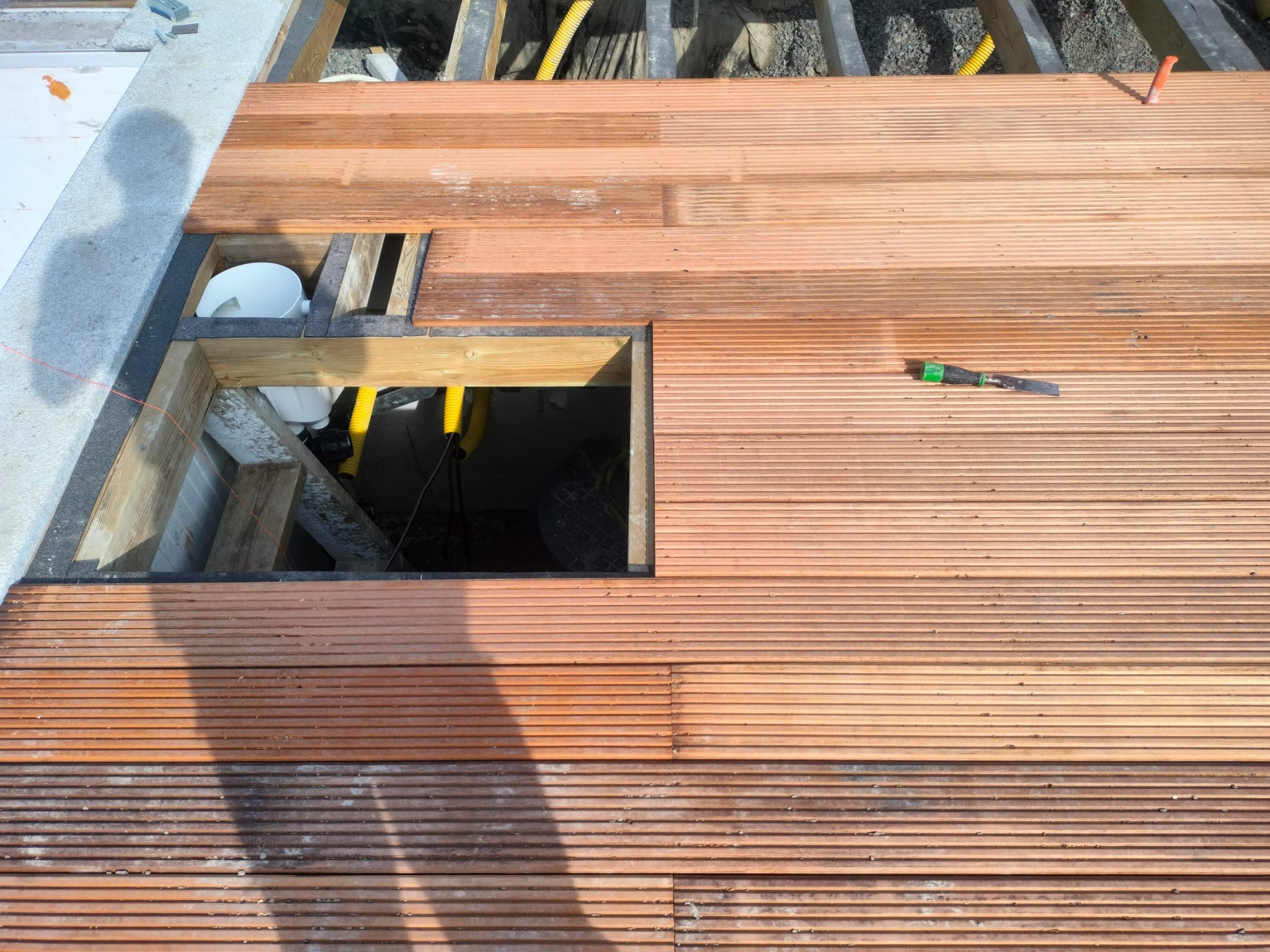 Montaż desek tarasowych w Sianożętach, widoczne elementy konstrukcyjne i instalacje pod tarasem. Deski ułożone w regularny wzór, częściowo zacienione.