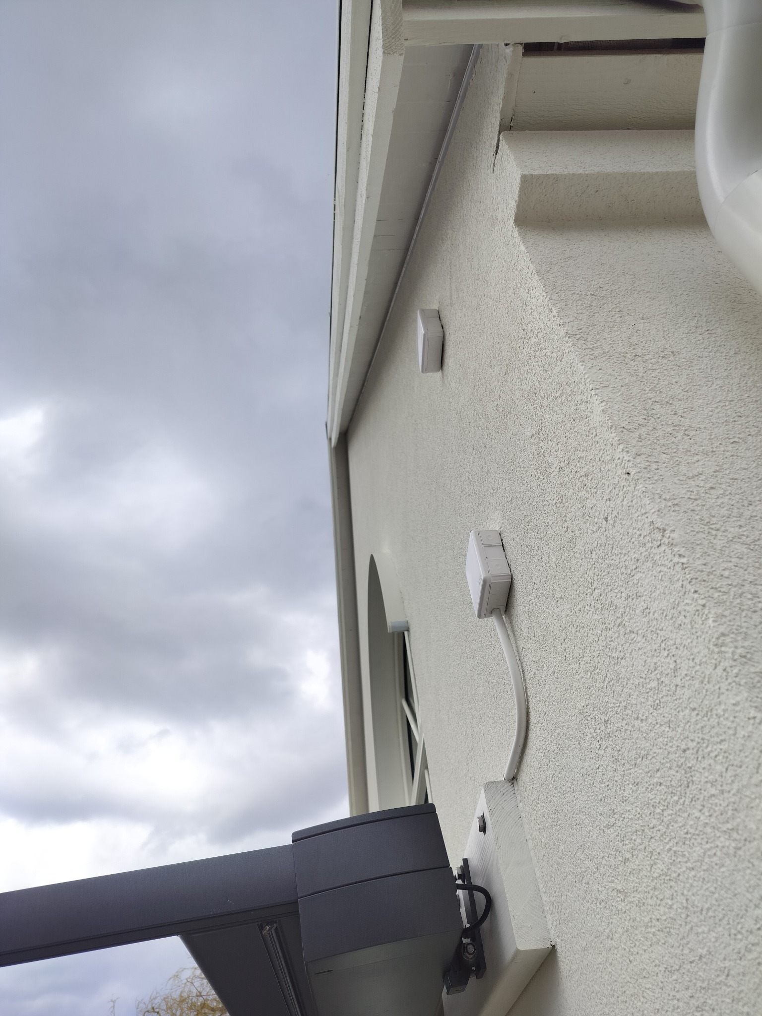 Elewacja budynku z widoczną pergolą w szarości, okno z kratami i instalacją elektryczną. Tło stanowi pochmurne niebo.