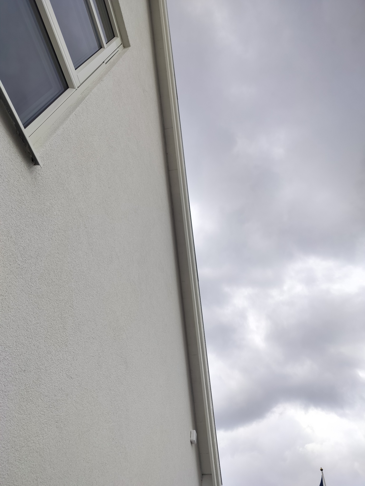 Fragment elewacji budynku z oknem, otynkowanej na biało, na tle pochmurnego nieba. Widoczny narożnik budynku i fragment rynny. Ujęcie z dołu.