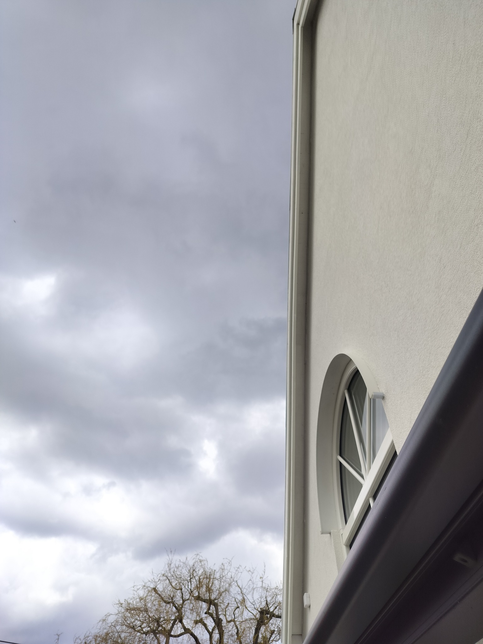 Fragment elewacji budynku z okrągłym oknem i widocznym niebem. Ujęcie z dołu podkreśla architekturę i specyficzny kształt okna. Minimalistyczny design budynku.