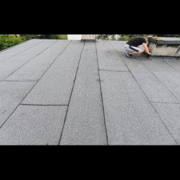 Henryk Hozakowski - Praca na dachu pokrytym szarą papą. Dekarz w trakcie uszczelniania komina. Widoczne narzędzia i fragmenty zieleni w tle.