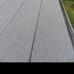 Szary, płaski dach pokryty papą termozgrzewalną, z widocznymi łączeniami. W tle fragment budynku i zielona trawa.