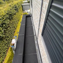 Henryk Hozakowski - Czarny, płaski dach po renowacji, widok z góry. Mur z jasnej cegły i zielony żywopłot w tle. Widoczny fragment drabiny oraz osoba pracująca na dachu.