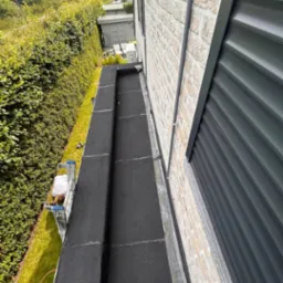 Czarny, płaski dach po renowacji, widok z góry. Mur z jasnej cegły i zielony żywopłot w tle. Widoczny fragment drabiny oraz osoba pracująca na dachu.