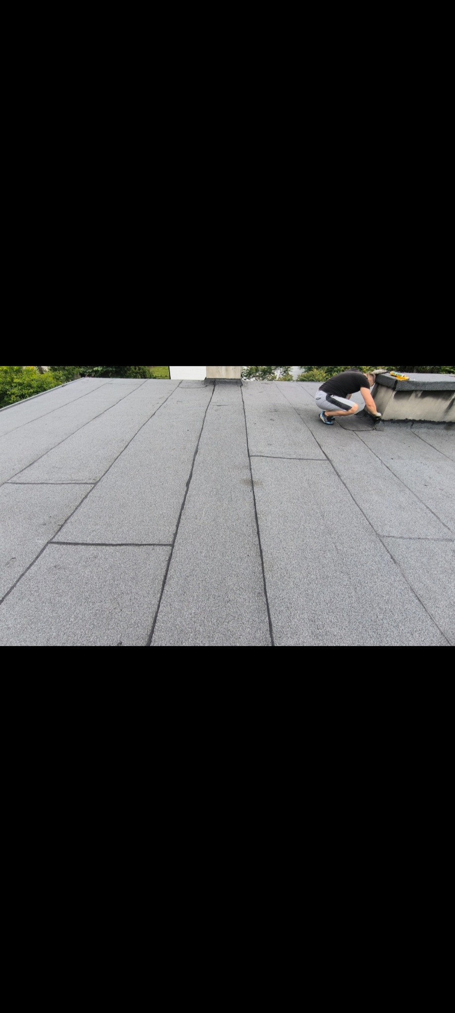 Praca na dachu pokrytym szarą papą. Dekarz w trakcie uszczelniania komina. Widoczne narzędzia i fragmenty zieleni w tle.