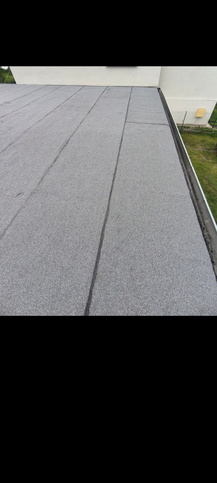 Szary, płaski dach pokryty papą termozgrzewalną, z widocznymi łączeniami. W tle fragment budynku i zielona trawa.