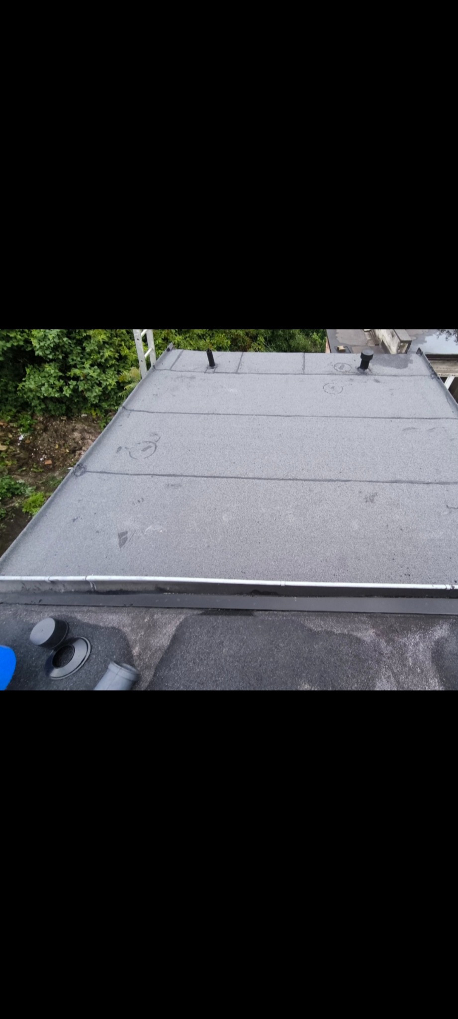 Widok z góry na płaski dach pokryty papą, z elementami instalacji wentylacyjnej i obróbkami blacharskimi. W tle zieleń drzew. Dach po renowacji.