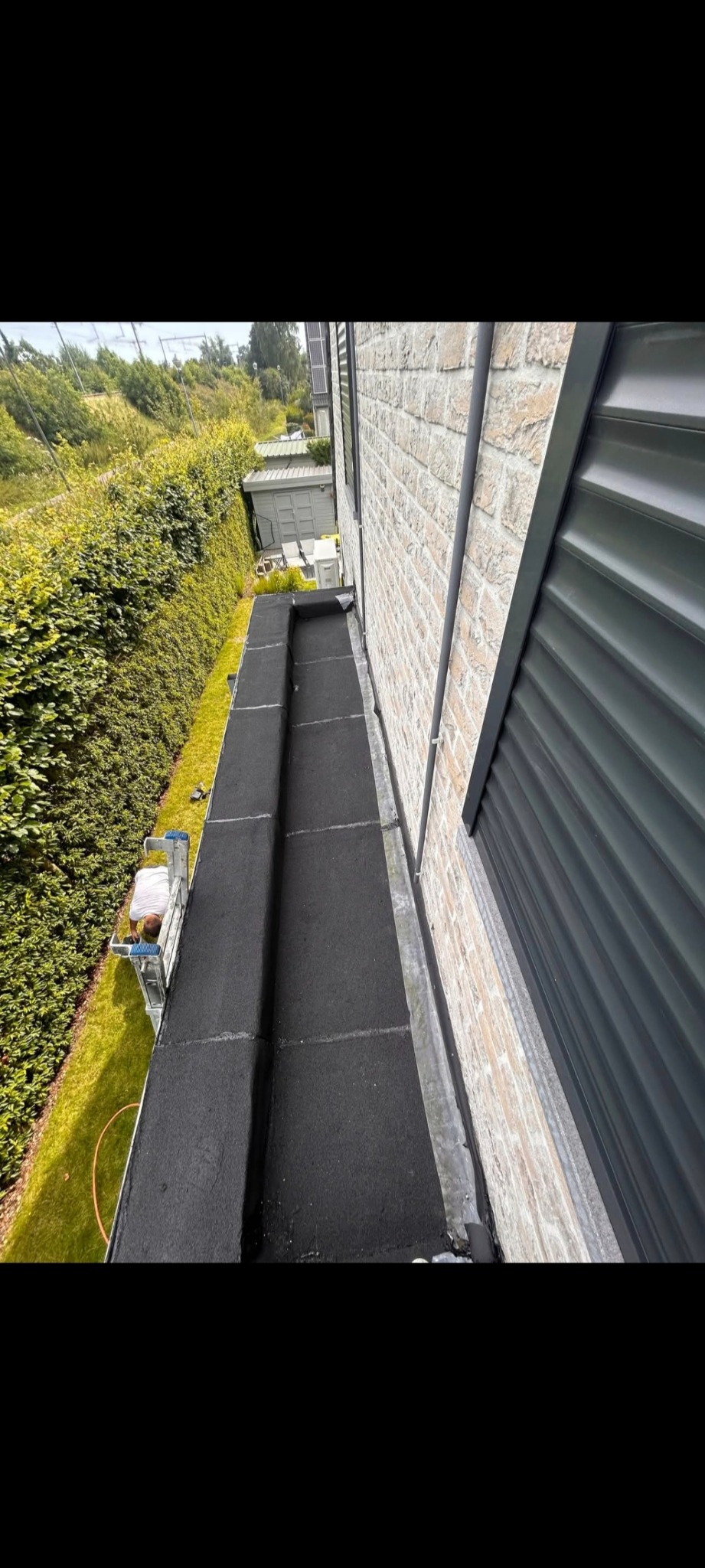 Czarny, płaski dach po renowacji, widok z góry. Mur z jasnej cegły i zielony żywopłot w tle. Widoczny fragment drabiny oraz osoba pracująca na dachu.