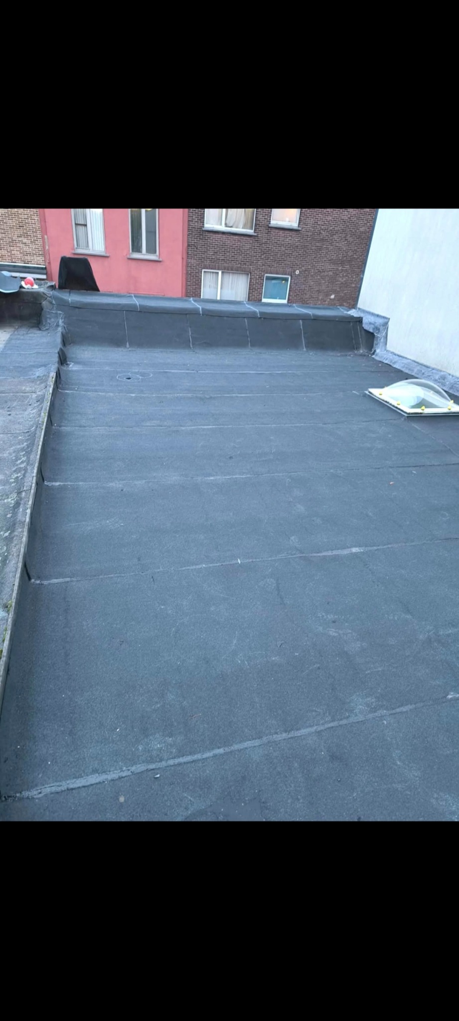 Wykończony płaski dach pokryty papą termozgrzewalną z widocznym świetlikiem dachowym. W tle budynki mieszkalne.