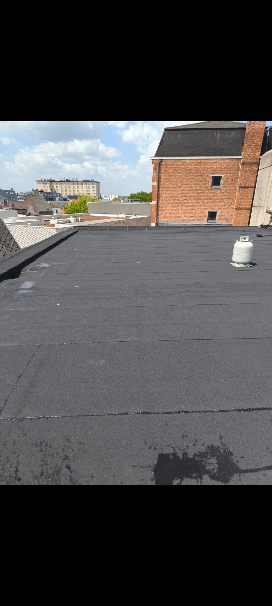 Czarny, płaski dach pokryty papą termozgrzewalną z widocznym budynkiem z czerwonej cegły w tle i butlą gazową. Widok z góry, słoneczny dzień.